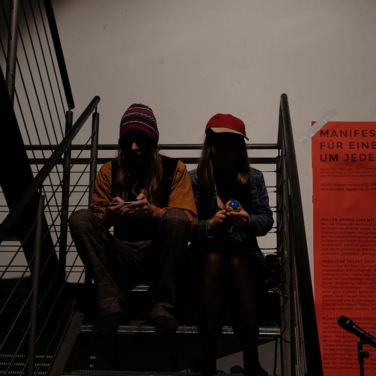 zwei Menschen sitzen auf einer Treppe, daneben ein großes rotes Plakat (Foto: SWR, SWR - Martin Horsky)