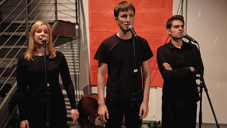 drei junge Menschen in schwarzer Kleidung sprechen gemeinsam in Mikrofone