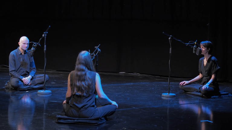 Drei Personen sitzen in einem abgedunkelten Raum vor Mikrofonen auf dem Boden. (Foto: SWR)