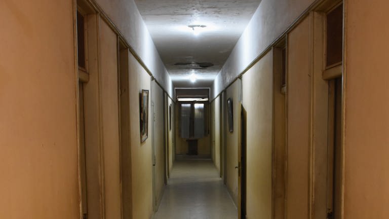 Langer, niedriger Gang, geschlossene Türen rechts und links, erleuchtet von einer einzelnen, hellen Glühbirne.