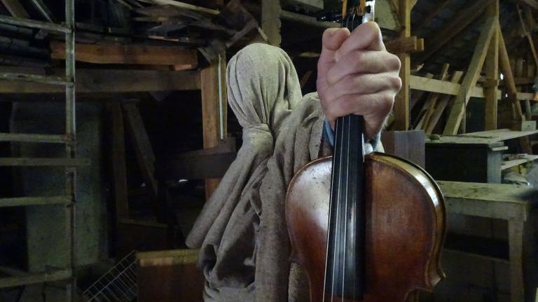 Eine in einem Sack verhüllte Person hält eine Violine in der Hand