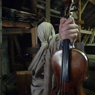 Eine in einem Sack verhüllte Person hält eine Violine in der Hand