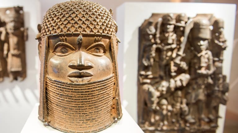 In einer Vitrine sind mehrere sogenannte „Benin-Bronzen“ auf weißem Untergrund ausgestellt. Im Vordergrund ist eine goldglänzende Büste eines geschmückten Kopfes. Im Hintergrund sind zwei Bronze-Relieftafeln an weißen Stellwänden aufgehängt.