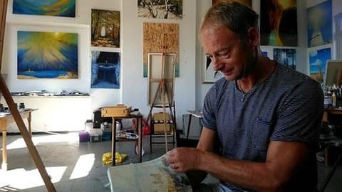 Der Künstler Armin Thommes in seinem Atelier in St. Goar, sitzend, im Hintergrund Staffelei und Bilder.