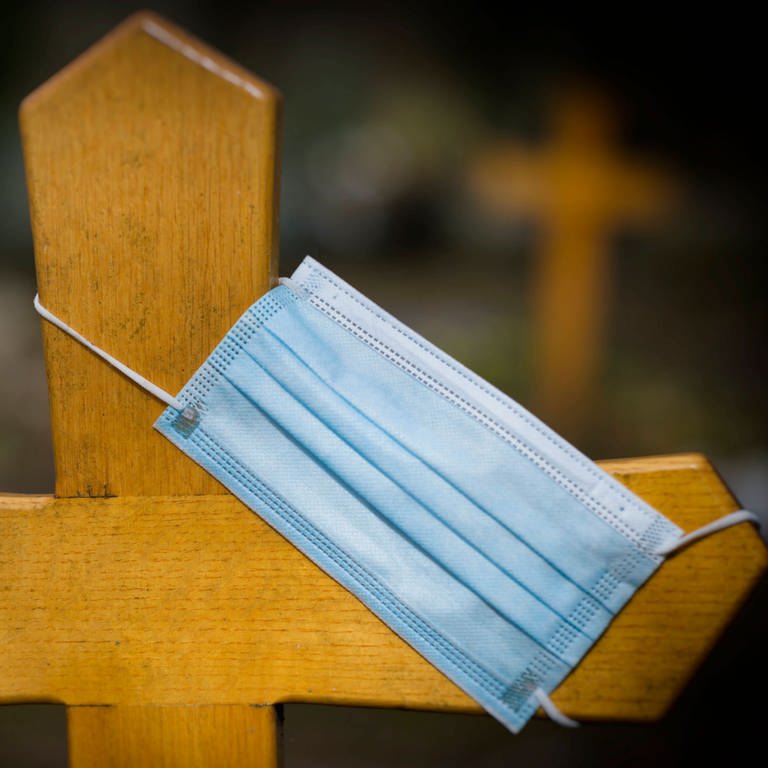 Eine Mundschutzmaske hängt an einem Grabkreuz (Foto: IMAGO, imago images/Michael Weber)