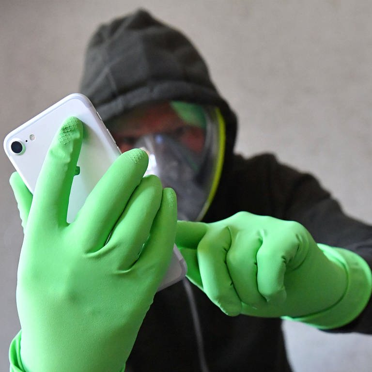 Ein Person hält ihr Smartphone mit Schutzhandschuhen in den Händen und trägt eine Vollgesichtsmaske (Foto: IMAGO, Frank Hoermann/Sven Simon via www.imago-images.de)