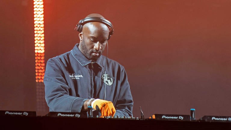Virgil Abloh: Ein schwarzer Mann in schwarzer Jacke an einem DJ-Pult. Er trägt einen Kopfhörer, die Bühne ist rot-gelb beleuchtet. (Foto: IMAGO, IMAGO / ZUMA Wire)
