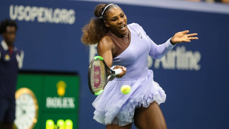 Eine schwarze Tennis-Spielerin, Serena Williams, im pastell-farbenen Dress. Ein Ärmel ist lang, der andere kurz und hautfarben. Sie schlägt mit dem Schläger in der rechten Hand einen Ball. Ihre Haare sind lang und lockig. (Foto: IMAGO, IMAGO / Shutterstock)
