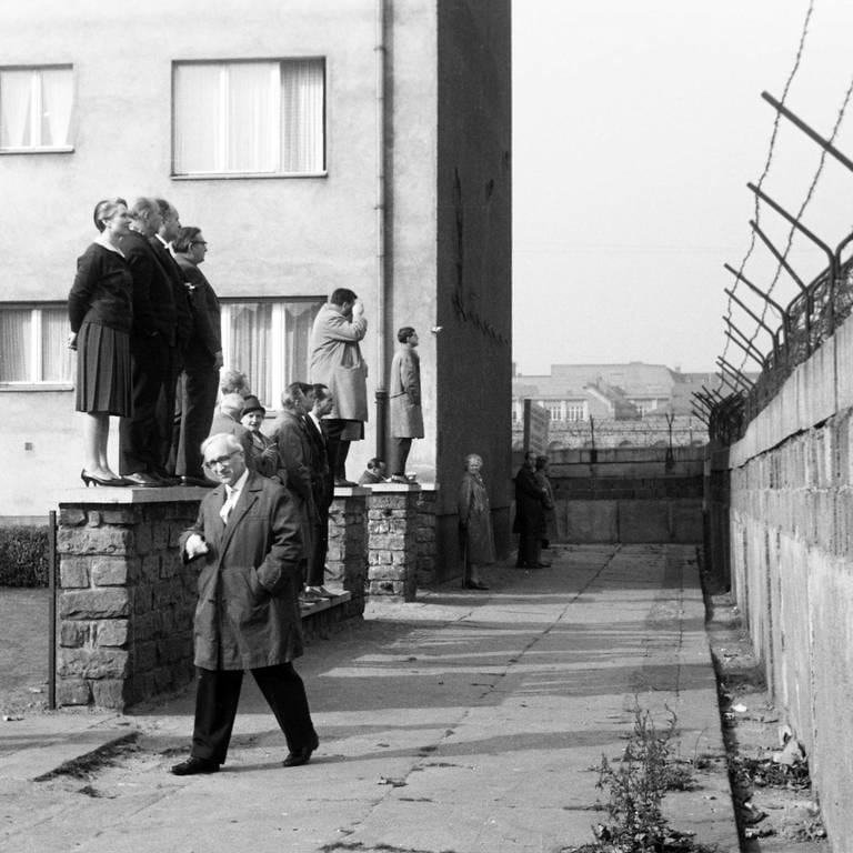 Eine Reisegruppe schaut von einer Vorgartenmauer über die Mauer hinweg vom Westteil in den Ostteil von Berlin, Deutschland 1962: Schwarz-weiß Bild, links stehen die Menschen, Männer und Frauen auf einem kleinen Mäuerchen, rechts befindet sich in mehreren Metern Abstand die Mauer, die oben mit Stacheldraht gesichert ist. (Foto: IMAGO, IMAGO / United Archives)