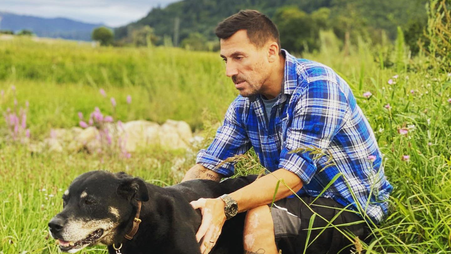 Christian Schindler sitzt auf einem großen Stein auf einer Wiese. Vor sich ein schwarzer Hund, den er streichelt. (Foto: Pressestelle, Annette Schindler)