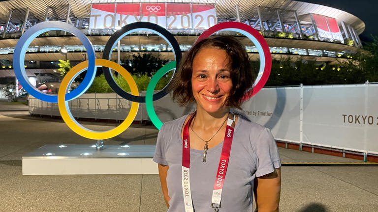 Kathrin Erdmann vor dem Nationalstation in Tokio. Hinter ihr die Olympischen Ringe in den Farben blau, gelb, scharz, grün und rot. Auf einem Banner am Stadion steht "Tokio 2020" geschrieben. (Foto: Kathrin Erdmann)