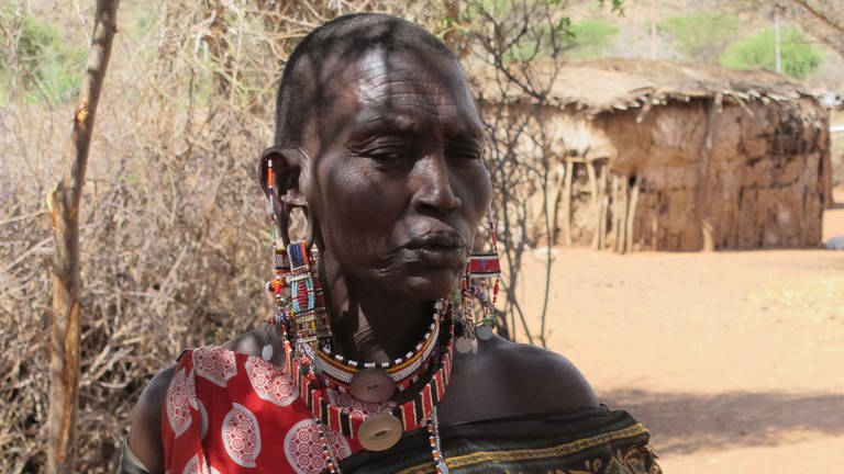 Stolze Massai-Frau, mit kahlem Schädel, bunten Tüchern um die Schulter und buntem Ohr- und Halsschmuck schaut den Betrachter mit skeptischem Blick an. Im Hintergrund eine Hütte. (Foto: Antje Diekhans)