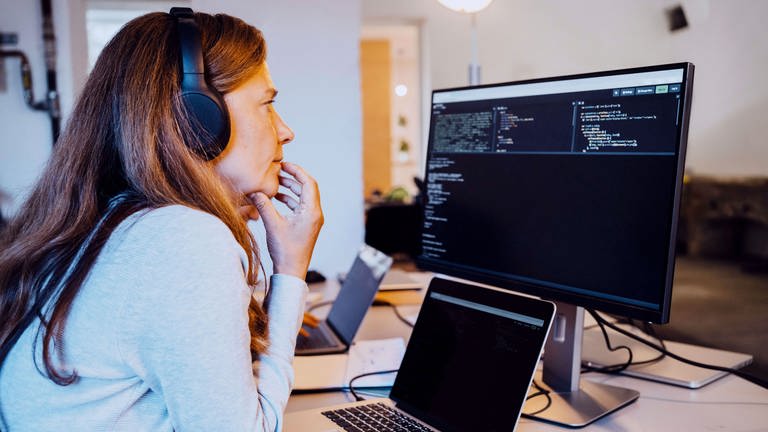 Eine Frau mittleren Alters mit schulterlangen Haaren und Kopfhörern sitzt vor einem Computer mit einer Programmier-Oberfläche und grübelt. Sie trägt einen grauen Pullover und stützt das Kinn mit der Hand ab. (Foto: IMAGO, IMAGO / MASKOT)