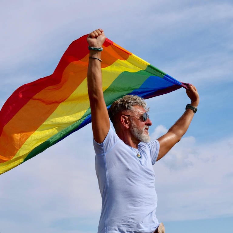 Portrait senior of a gray-haired elderly Caucasian man with a beard and sunglasses holding a rainbow LGBTQIA flag against a sky (Foto: picture-alliance / Reportdienste, picture alliance / Zoonar | Oksana Fedorchuk)