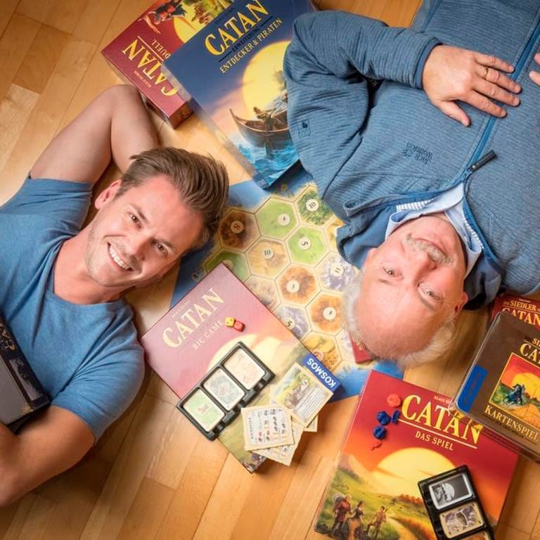 Benjamin (li.) und Klaus Teuber (re.), Spieleentwickler, diagonal zueinander auf dem Boden liegend, umgeben von Spielekartons mit der Aufschrift "Catan" (Foto: Patrick Liste)