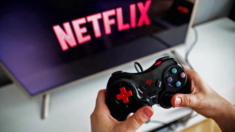 Netflix will sich in Zukunft verstärkt um den Ausbau seiner Gaming-Abteilung kümmern - Hände halten den Controller einer Spielekonsole vor einem TV-Bildschirm auf dem das Netflix-Logo angezeigt wird. (Foto: IMAGO, IMAGO / NurPhoto)