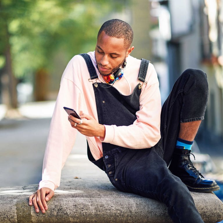 Unsere SWR2 Netztipps für den Sommer 2021: ein junger, schwarzer Mann sitzt mit aufgestütztem Bein auf einer Mauer, an einem sonnigen Tag. Im Hintergrund ist ein Platz in einer Stadt mit Bäumen gesäumt zu sehen. Der junge Mann trägt einen hellrosa Pullover und eine schwarze 78 Latzhose. Er hat einen Kopfhörer um den Hals gehängt und scrollt in seinem Smartphone. (Foto: IMAGO, IMAGO / ingimage)