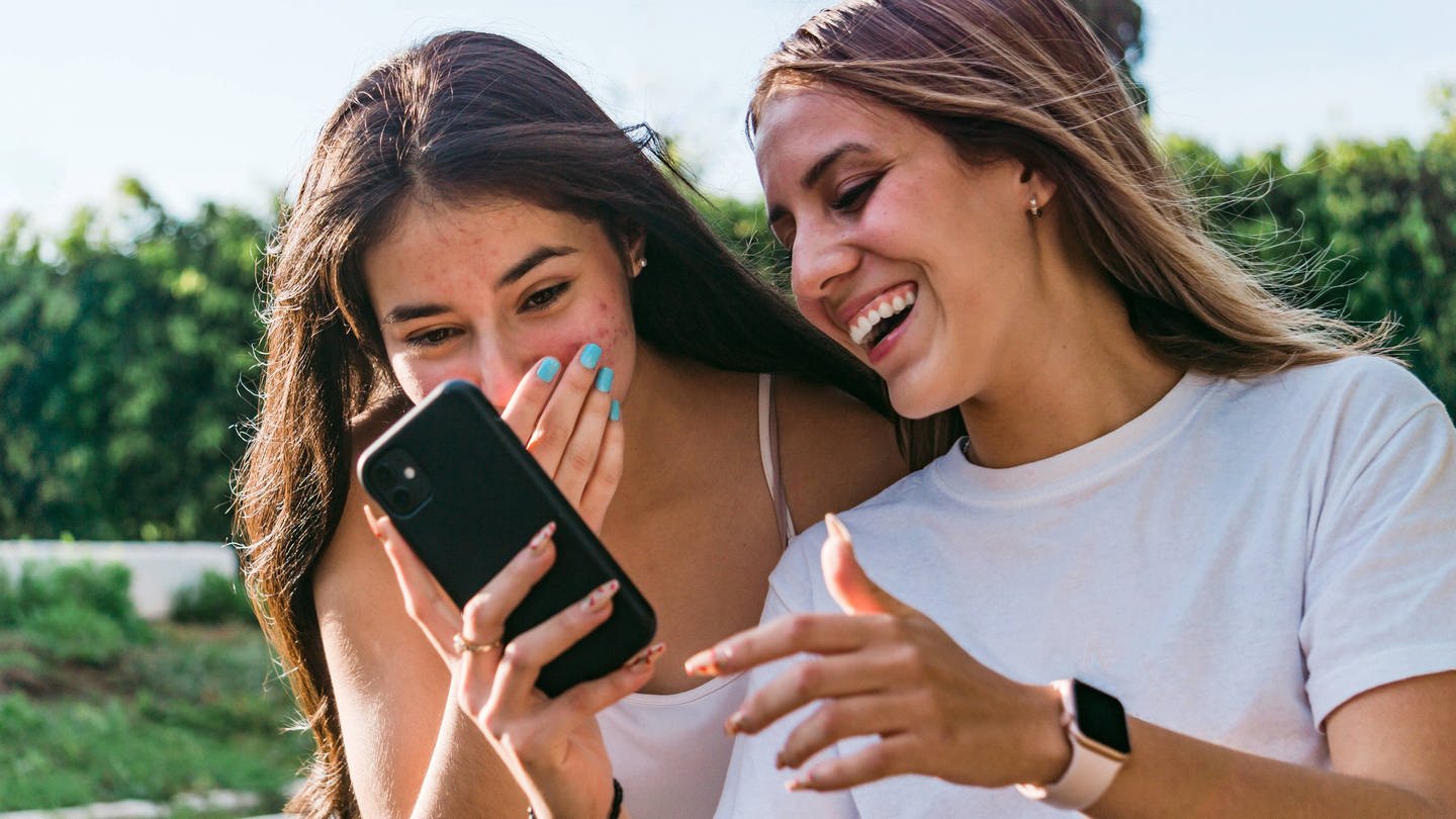 Zwei weiße Teenie-Mädchen mit längeren Haaren blicken auf ein Smartphone und amüsieren sich. (Foto: IMAGO, IMAGO / Addictive Stock)