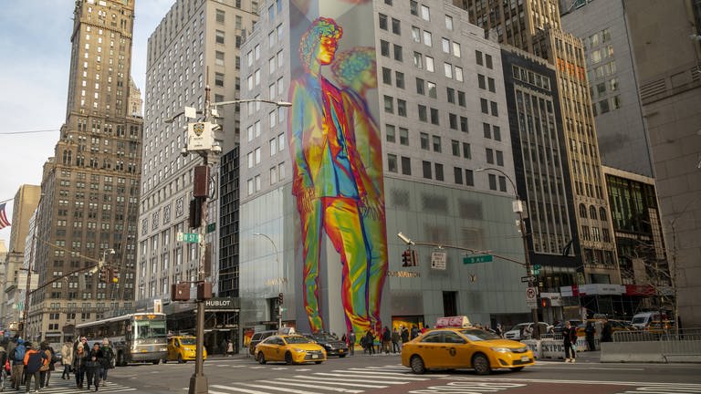 Eine Hausecke an einem grauen Gebäude in New York City, davor eine Straßenkreuzung. An der Hausecke ist eine holografisch anmutende, vielfarbige Installation, die einen Mann im Anzug zeigt. (Foto: IMAGO, IMAGO / Levine-Roberts)