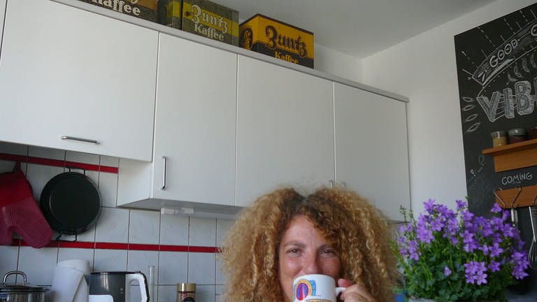 Ruthe trinkt Kaffee aus einer „Zuntz“-Kanne, im Hintergrund die Zuntz-Kaffebehälter (Foto: Igal Avidan)
