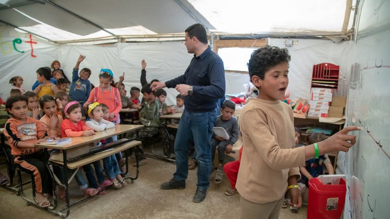 Kinder sitzen auf Schulbänken, hören dem Lehrer zu und schauen auf einen Jungen der an der Tafel steht (Foto: Zeltschule)