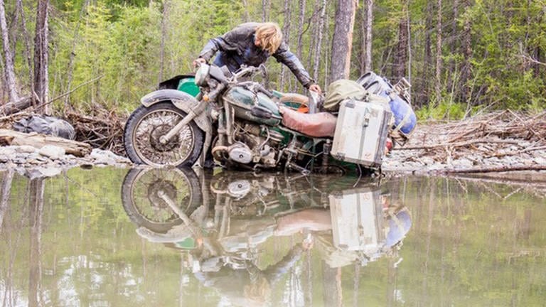 Das Hinterrad eines vollgepackten Motorrads ist im Sumpf eingesunken. Der Fahrer versucht es herauszuziehen.