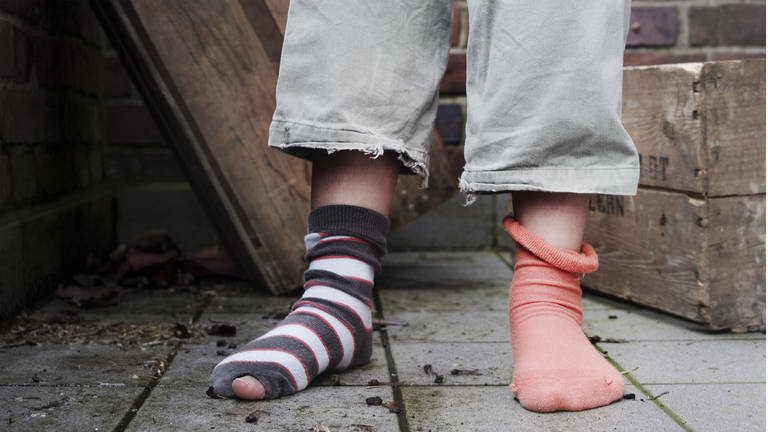 Ein Kind steht in abgetragener Kleidung ohne Schuhe und mit zwei verschiedenen Socken in einem Hinterhof