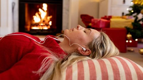Weihnachtstipps: Entspannte Frau mit Kopfhörern am Kamin (Foto: IMAGO, Wavebreak Media Ltd)