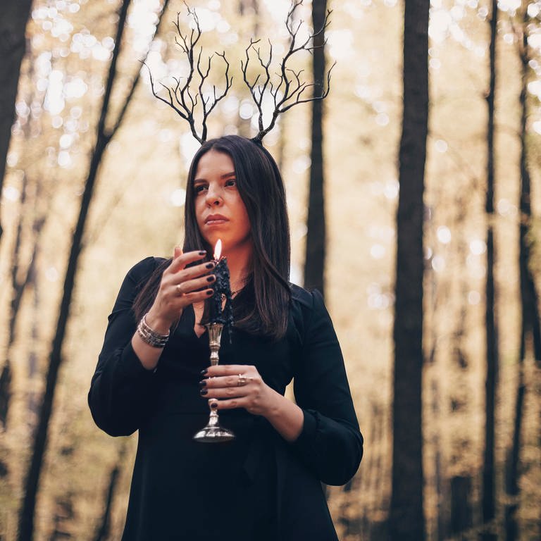 Eine Frau im dunklen Look mit brennender Kerze im Wald