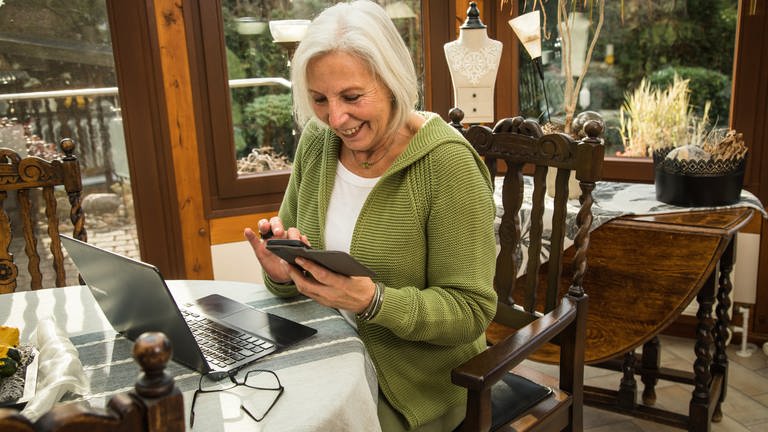 Eine Rentnerin schaut in ihr Smartphone während sie am Laptop arbeitet