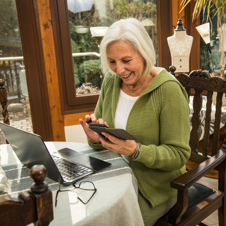 Eine Rentnerin schaut in ihr Smartphone während sie am Laptop arbeitet (Foto: picture-alliance / Reportdienste, picture alliance / dpa-tmn | Christin Klose)