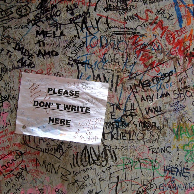 Namen und Schrift, Gekritzel an einer Wand. Darauf ist ein Papierschild geklebt mit der Aufschrift "Please don´t write here" 