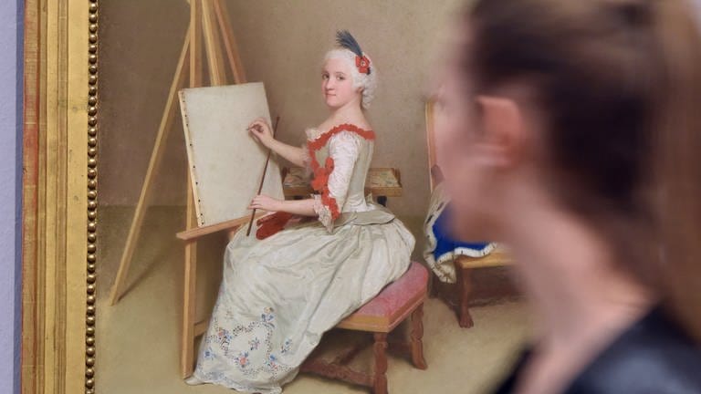 Ein Gemälde eines Mädchens an der Staffelei hängt in einem Raum, eine Frau schaut es sich im Vorrübergehen an 