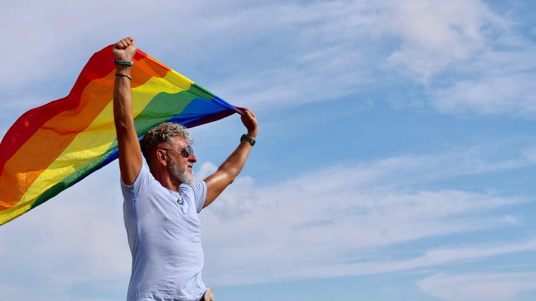 Portrait senior of a gray-haired elderly Caucasian man with a beard and sunglasses holding a rainbow LGBTQIA flag against a sky