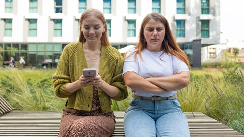 Zwei Frauen sitzen nebeneinander auf einer Bank, eine schaut auf ihr Handy, die andere scheint genervt zu sein (Foto: IMAGO, IMAGO/Pond5 Images/xarucikx)