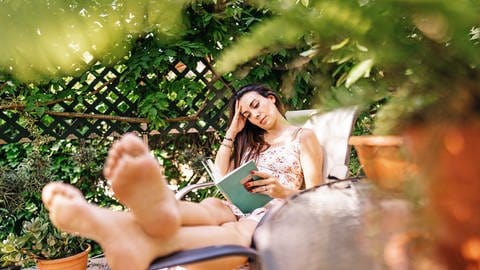 Eine junge, schwangere, schwarzhaarige Frau liegt auf einem Liegestuhl und liest in einem grünen Buch. Sie trägt ein beiges langes Kleid mit Blumenmuster und stützt ihren Kopf in der rechten Hand ab. Sie ist von vielen grünen Pflanzen umgeben. Ihre Fußsohlen sind unscharf im Vordergrund zu sehen. (Foto: IMAGO, IMAGO / Addictive Stock)