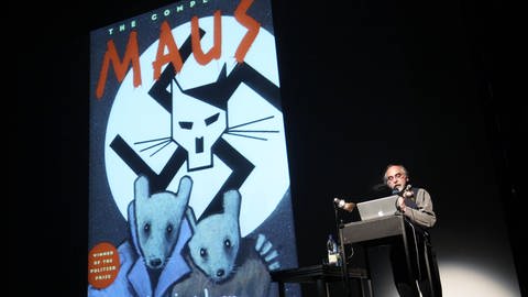 Bild eines älteren Mannes an einem Rednerpult, neben ihm projiziert das Buchcover von 'Maus'. Es zeigt einen Art stilisierten Hitler als Maus in einem Hakenkreuz. (Foto: IMAGO, IMAGO / BRIGANI-ART)
