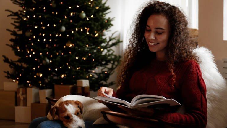 Eine glückliche junge Frau sitzt neben einem Hund lesend vor dem Weihnachtsbaum