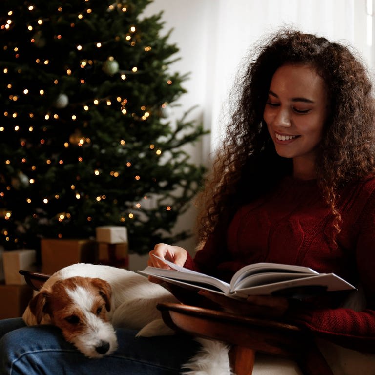 Eine glückliche junge Frau sitzt neben einem Hund lesend vor dem Weihnachtsbaum (Foto: IMAGO, IMAGO / Westend61)