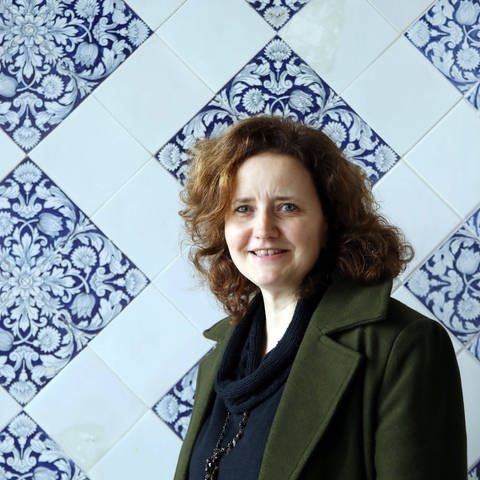 Autorin Julia Schoch in einem olivgrünen Blazer vor einer blau-weiß gekachelten Wand.
