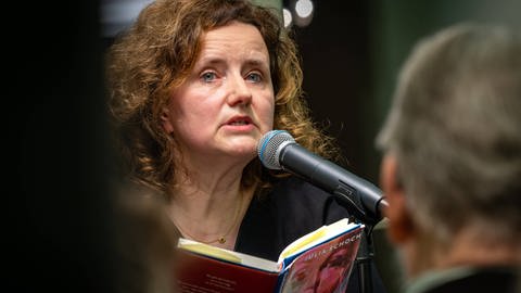 Julia Schoch am Mikrofon. Sie hält ihr Buch "Das Liebespaar des Jahrhunderts" in der Hand, aus dem sie liest.