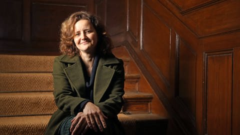 Autorin Julia Schoch in einer olivgrünen Jacke. Sie sitzt auf einer Treppe in einem holzgetäfelten Treppenhaus.  (Foto: IMAGO, IMAGO / Sabine Gudath)