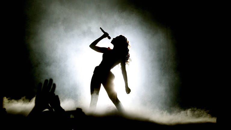 Beyoncé Knowles in der Berliner O2 World: Die Silhouette einer langhaarigen Frau auf einer benebelten Bühne, sie lehnt sich leicht seitlich im breitbeinigen Stand und hält ein Mikrofon vor ihre Lippen. (Foto: IMAGO, IMAGO / BRIGANI-ART)