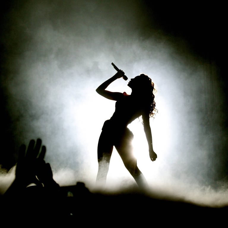Beyoncé Knowles in der Berliner O2 World: Die Silhouette einer langhaarigen Frau auf einer benebelten Bühne, sie lehnt sich leicht seitlich im breitbeinigen Stand und hält ein Mikrofon vor ihre Lippen. (Foto: IMAGO, IMAGO / BRIGANI-ART)
