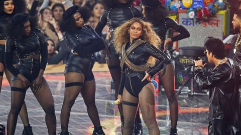 Beyoncé Knowles und Bruno Mars beim Superbowl 50 20152016, während der Halbzeitspausen-Show: Auf einer Bühne stehen mehrere in Leder gekleidete afroamerikanische Frauen und Männer, rechts zwei Männer, die zu einer Frau in die Mitte blicken, die Frau hat blondierte, lange Haare und trägt einen Body zu ihrer Lederjacke, sowie gekreuzte Munitionsgurte über die Brust. Hinter ihr stehen mehrere Frauen in einem ähnlichen Outfit, mit schwarzen Bérets.