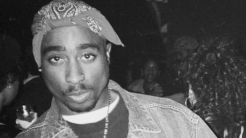 Der Rapper und Schauspieler Tupac Shakur: schwarz-weiß Bild eines afroamerikanischen Mannes in Jeansjacke und Adidas-Pullover. Er zeigt beide Mittelfinger in die Kamera. Er trägt auf dem Kopf ein Bandana und hat einen Oberlippenbart. (Foto: IMAGO, IMAGO / MediaPunch)