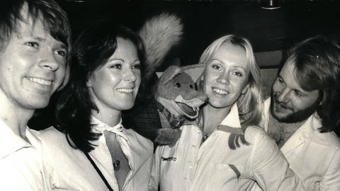 ABBA lachen in die Kamera, eine Stoff-Handpuppe zwischen Agnetha Fältskog und Anni-Frid Lyngstad 