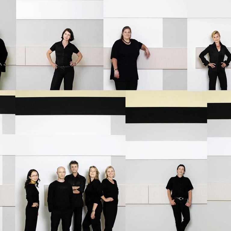 Mitglieder des SWR Vokalensembles Stuttgart  stehen einzeln oder in Grüppchen vor einer hellen Wand