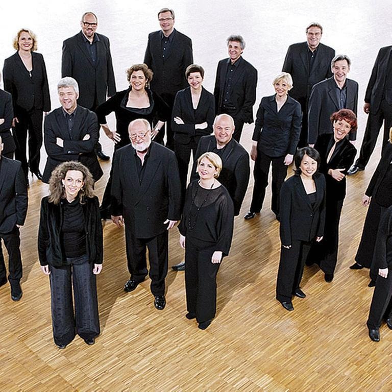 Die Sängerinnen und Sänger vom SWR Vokalensemble Stuttgart stehen, schwarz gekleidet, in einer lockeren Gruppe auf einem Parkettboden