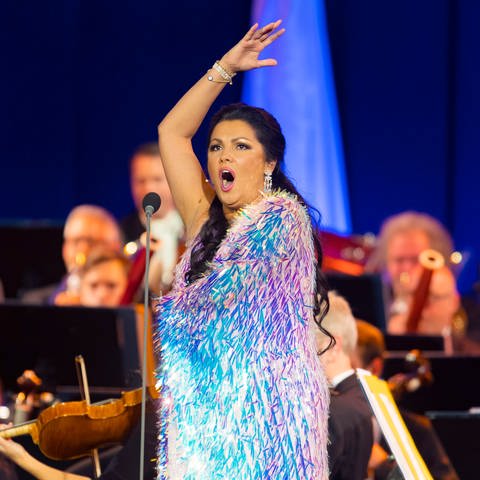 Anna Netrebko singend auf der Bühne. Ihr Kleid schimmert in verschiedenen Blau-Violetttönen